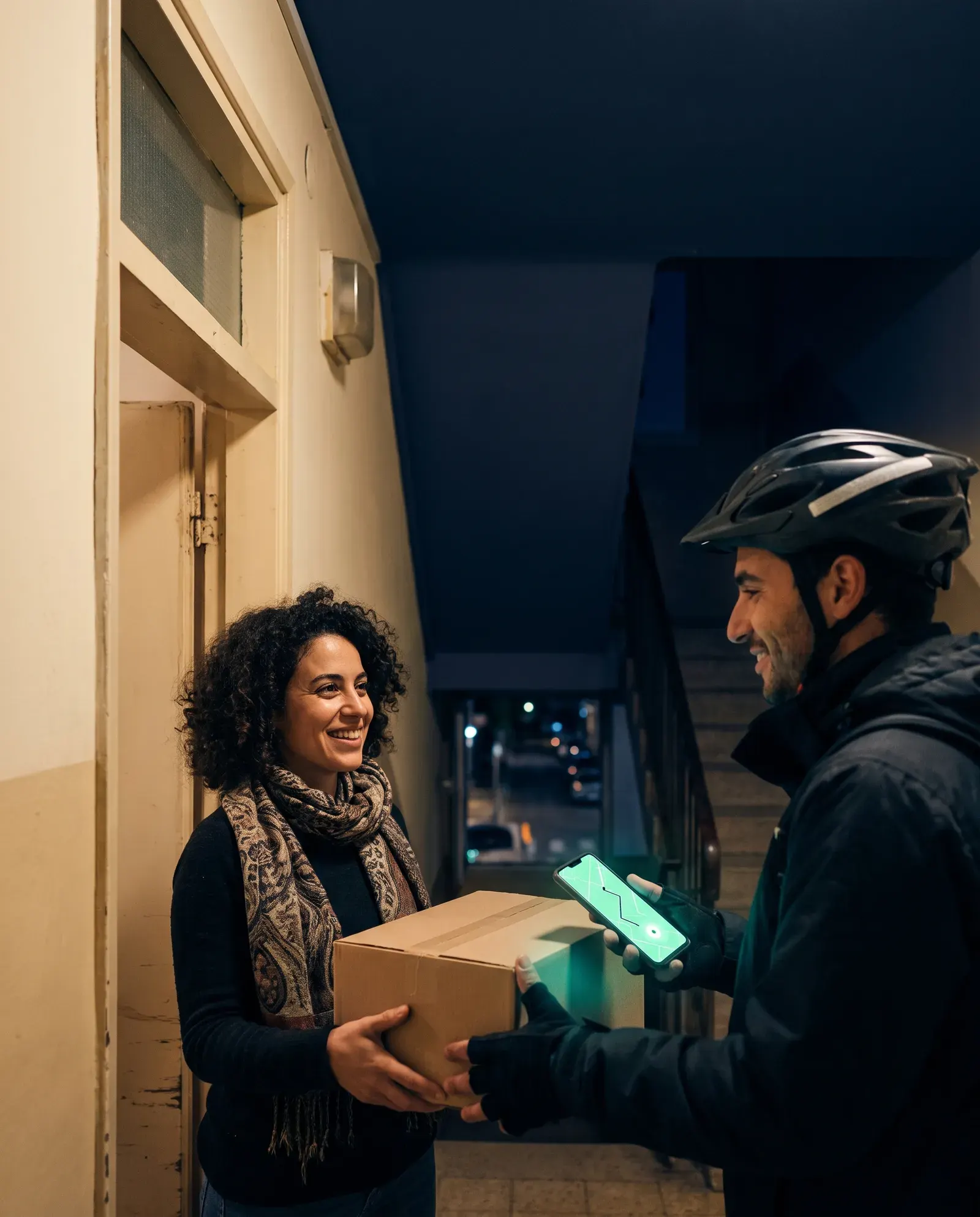 שליח עם קסדה מוסר חבילה לאישה מחייכת בפתח הבניין, וטלפון זוהר בירוק בידו